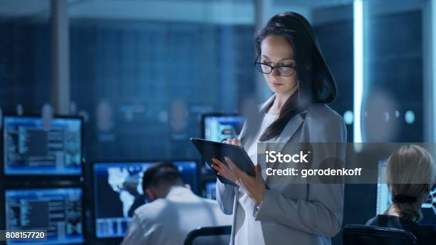 Young Female Engineer Uses Tablet In System Control Center In The Background Her Coworkers Are At Their Workspaces With Many Displays Showing Valuable Data Stock Photo - Download Image Now