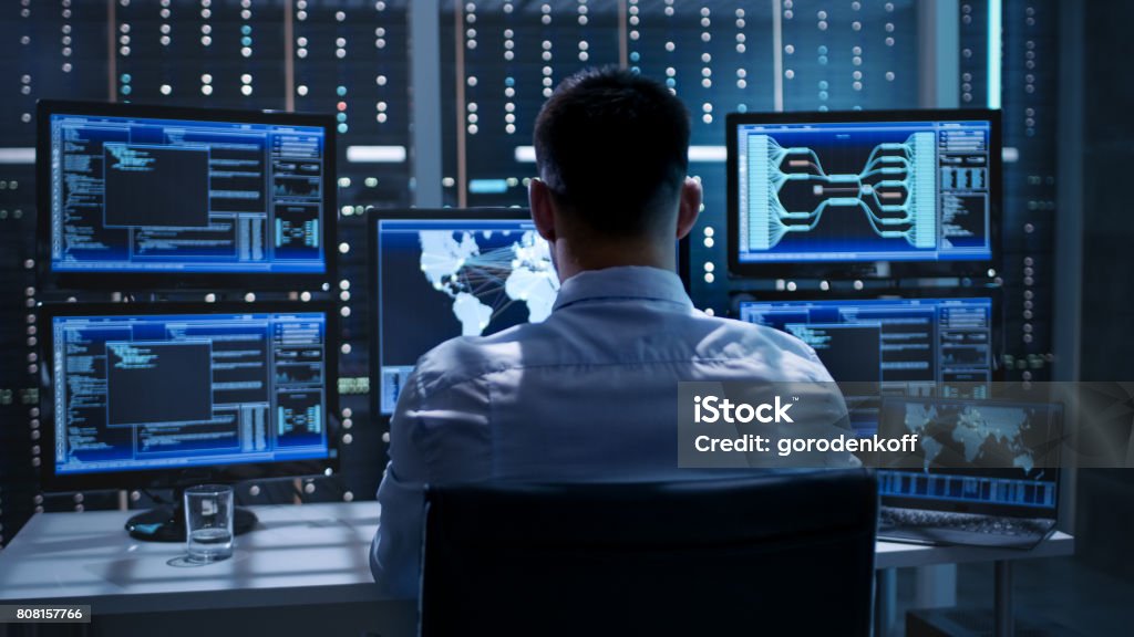 System Security Specialist Working at System Control Center. Room is Full of Screens Displaying Various Information. Network Security Stock Photo