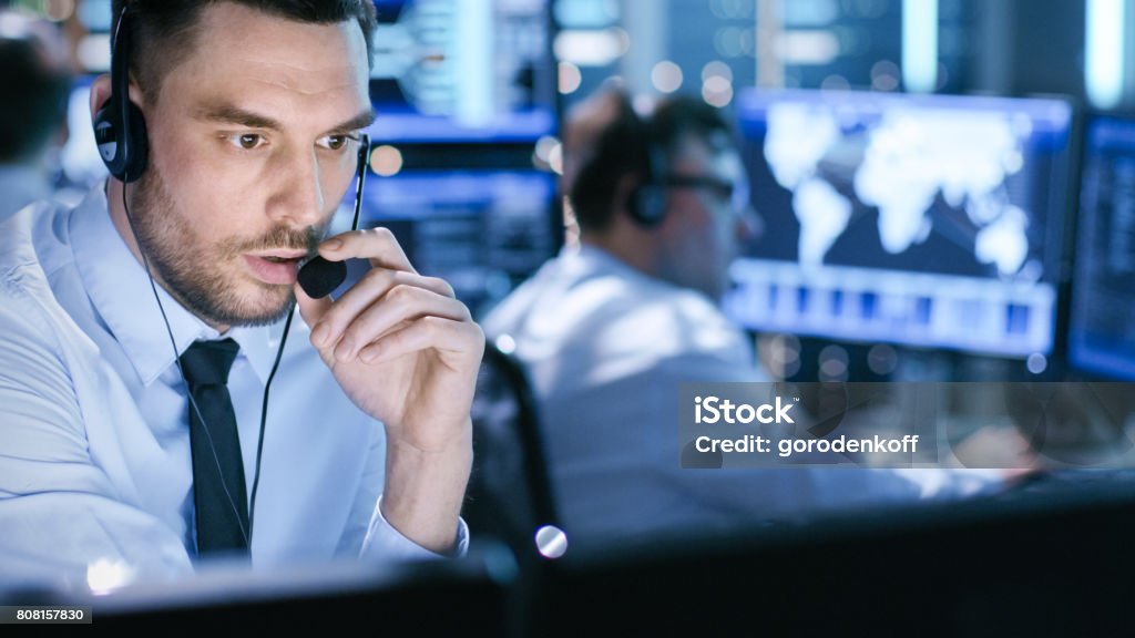 In Monitoring Room Technical Support Specialist Speaks into Headset. His Colleagues are Working in the Background. Security Stock Photo