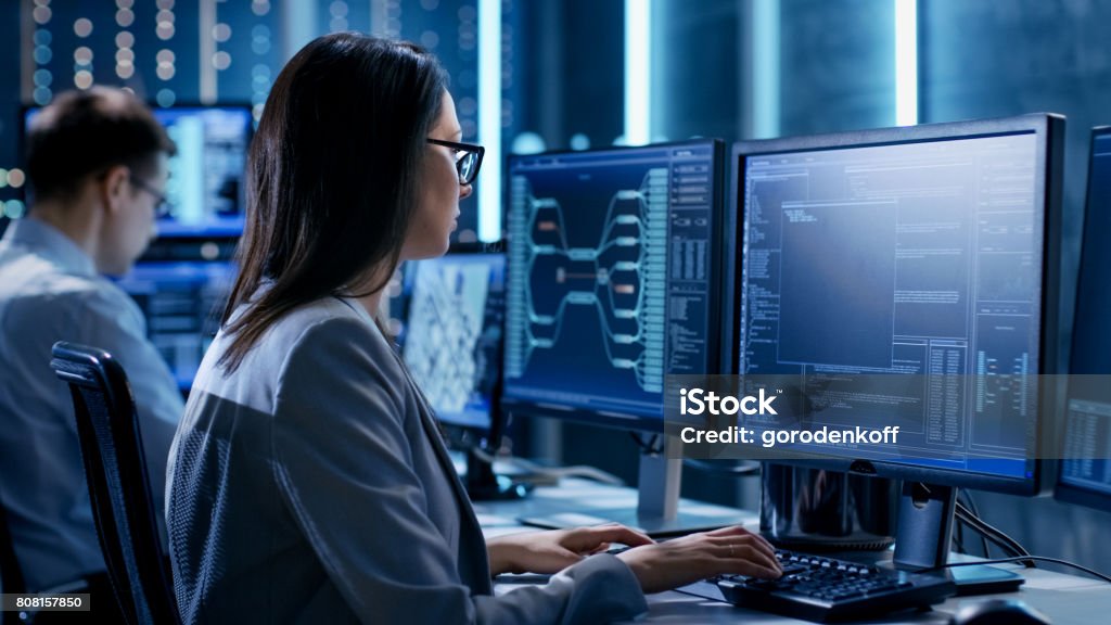 Over The Shoulder Footage of Female IT Engineer Working in Monitoring Room. She Works with Multiple Displays. Control Room Stock Photo