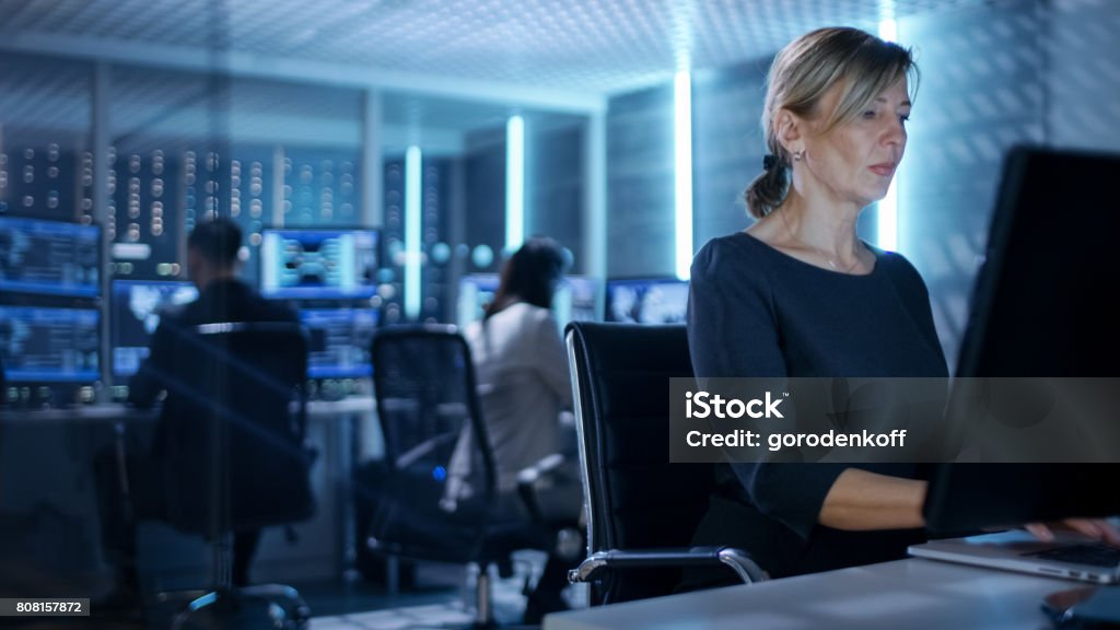 Female IT Engineer Works on Her Desktop Computer in Government Surveillance Agency. In the Background People at Their Workstations with Multiple Screens Showing Graphics. Police Force Stock Photo