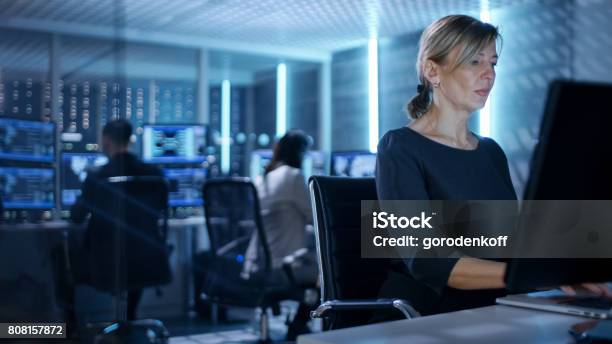 Female It Engineer Works On Her Desktop Computer In Government Surveillance Agency In The Background People At Their Workstations With Multiple Screens Showing Graphics Stock Photo - Download Image Now