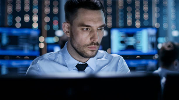Close-up of a Professional Technical Controller Sitting at His Desk with Multiple Displays Before Him. In the Background His Colleagues Working in System Control Center. Close-up of a Professional Technical Controller Sitting at His Desk with Multiple Displays Before Him. In the Background His Colleagues Working in System Control Center. facilities protection services stock pictures, royalty-free photos & images