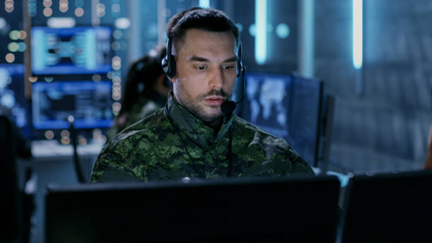 istock Close-up shot of Military Technical Support Professional Gives Instructions Using Headset. He's in a Monitoring Room with Other Officers and Many Working Displays in Background. 808157946