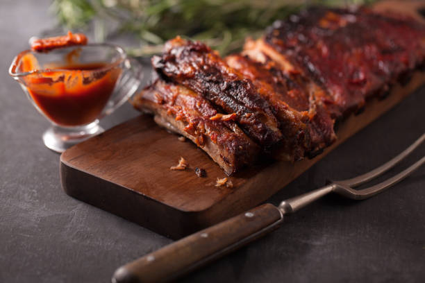 Delicious barbecued ribs seasoned with a spicy basting sauce and served with chopped fresh vegetables on an old rustic wooden chopping board in a country kitchen stock photo