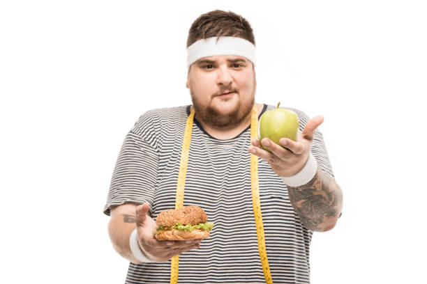 jeune homme joufflu, regardant la caméra tout en maintenant le burger et apple isolé sur blanc - food measuring hamburger dieting photos et images de collection