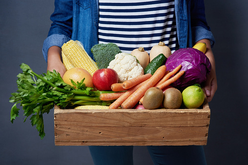 Studio shot of an unrecognizable woman holding a wooden crate full of fruit and vegetables against a blue background