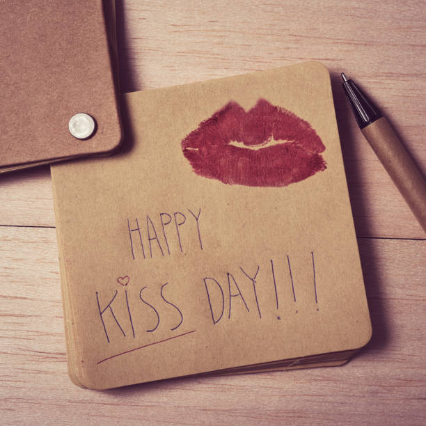 high-angle shot of a brown note with the text happy kiss day written in it and the mark of a pair of red lips, on a wooden table next to a pen