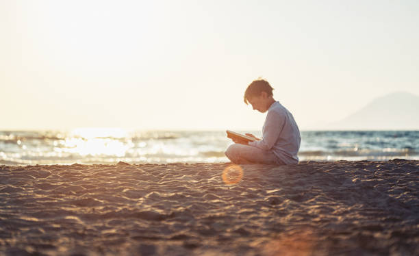 istock Cute boy reading a book on beach at sunset 808424896