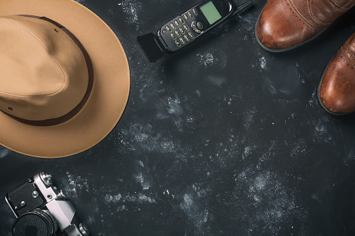 Tourism and travel concept. Vintage film camera, brown shoes, fedora hat and old mobile phone on black stone background. Free space for your text.