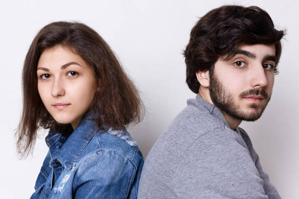 istock Two young people sitting back to back looking directly into camera. A young couple: brunette girl in jean clothes and stylish bearded man in grey sweater sitting backs relaxing 808482092