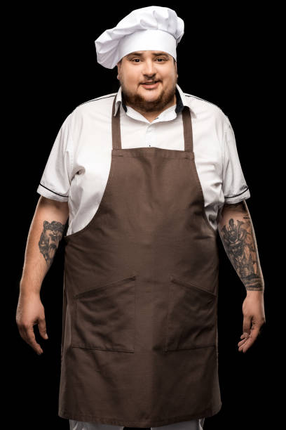 istock tattooed smiling young chef standing in white hat and apron isolated on black 808515202