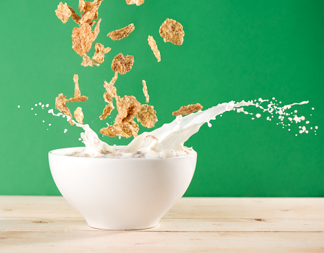 Corn Flakes dropping into bowl causing milk splash over wooden table on green background