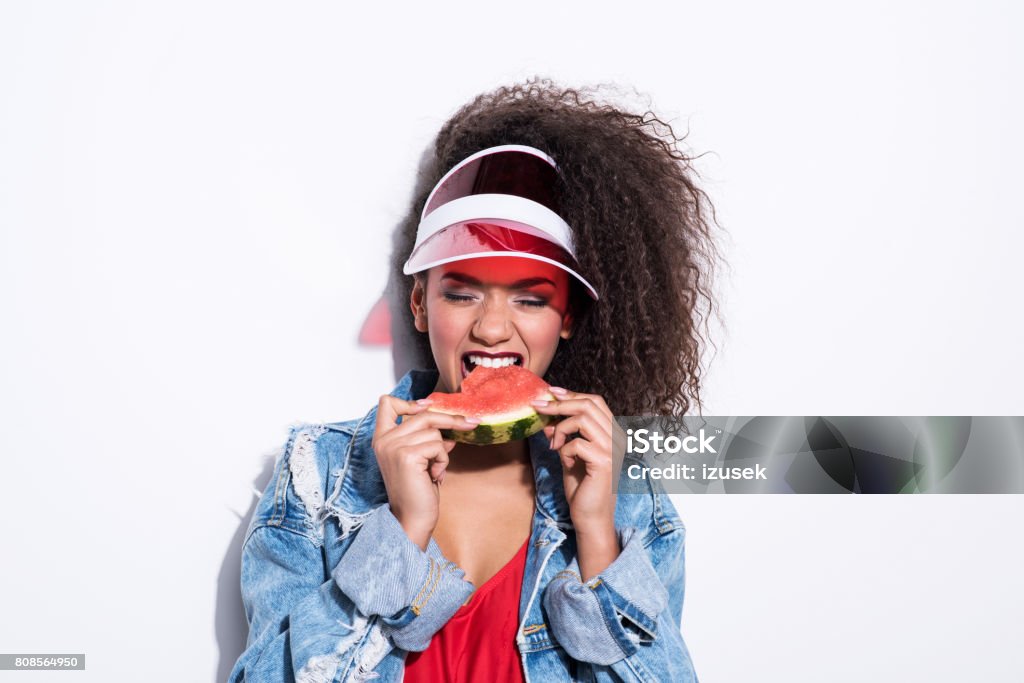 80's style young summer woman eating watermelon Studio portrait of 80’s style young woman wearing oversized worn denim jacket and eating watermelon. White background. Watermelon Stock Photo