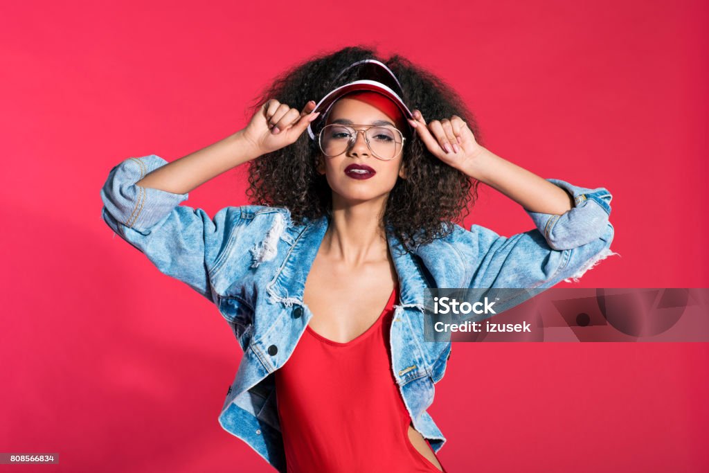 Fashion portrait of 80's style young woman weraign worn denim jacket Fashion portrait of 80’s style happy young woman wearing oversized worn denim jacket and glasses. Red background. 1980-1989 Stock Photo