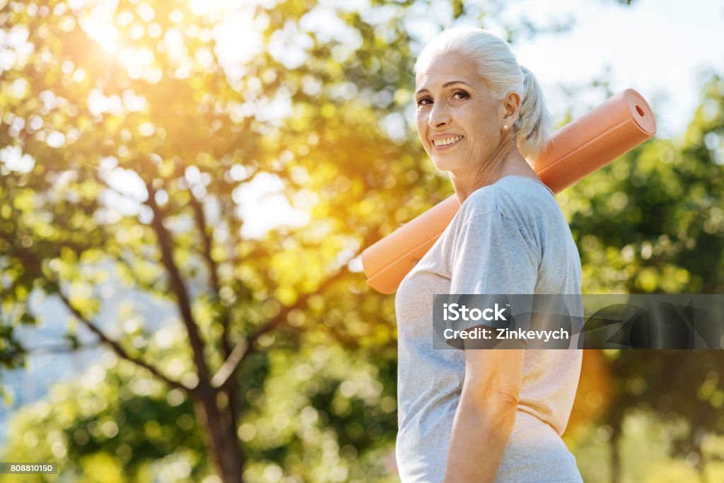 Cheerful senior woman going to enjoy yoga in the park Stay active. Cheerful smiling aged woman holding yoga carpet on the shoulder and looking in the camera while standing in the park Yoga Stock Photo