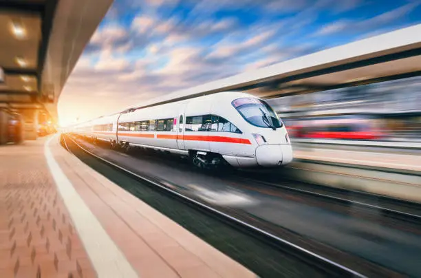 Photo of White modern speed train in motion on railway station at sunset. Train on railroad track with motion blur effect in Europe in evening. Railway platform. Industrial landscape. Railway tourism