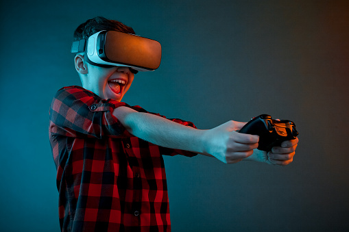 Laughing kid in checkered shirt playing gamepad while wearing VR goggles on studio background.