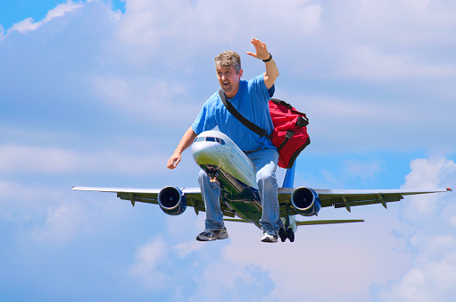 A happy man with a red duffel bag is waving as he flies through the air riding on an airplane like someone would ride a horse - happy frequent flyer, flying traveler and travel agency concept.