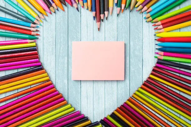 Top view of colored pencils shaped heart symbol with a blank note paper