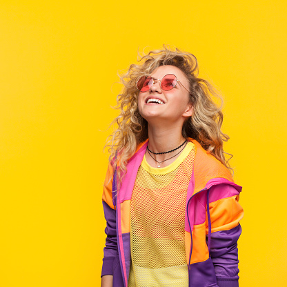 Smiling blonde curly haired girl wearing sunglasses, sheer shirt and jacket with zipper looking up.