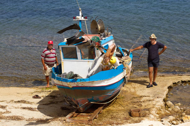Sampieri, Ragusa Province, Sicily: Two fishermen wait next to their blue and white fishing boat for an extra hand to help launch it into the Mediterranean Sea in Sampieri, Sicily.