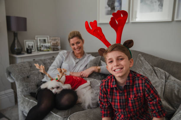 Happy little boy is smiling for the camera while wearing christmas antlers. His mother is sitting next to him with their pet dog on her lap.