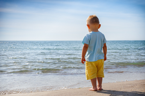 day barefoot boy 1.5 years in yellow shorts and a blue shirt stands on the beach near the water on the sand and looks at the approaching wave