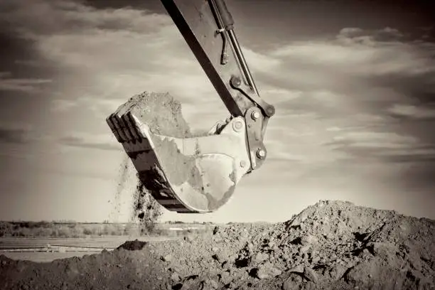Excavator arm and scoop full of dirt at construction site against sky, black and white, sepia toned image