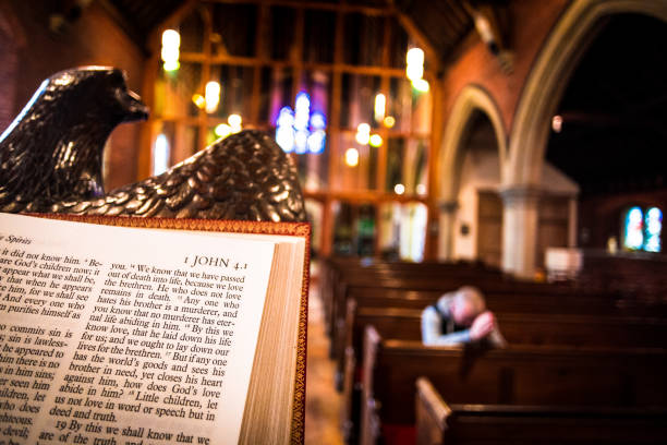 Man praying on pew inside English Anglican church with focus on bible in foreground Close up image of the Holy Bible open at the book of John on the altar of an Anglican church. The pews and stained glass windows of the interior of the church are blurred out of focus in the background, and warm orange sunlight is streaming in through the windows. On one of the front pews a young man is bowed in prayer, and he too is blurred out of focus. Horizontal colour image with copy space. anglican eucharist stock pictures, royalty-free photos & images