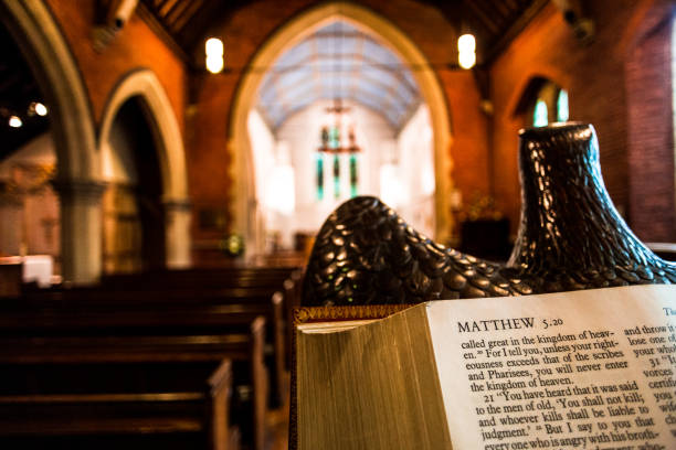 Open bible on the Lectern inside an English Anglican church Close up image of the Holy Bible open at the book of Matthew on a lectern inside an Anglican church. The pews and stained glass windows of the interior of the church are blurred out of focus in the background, and warm orange sunlight is streaming in through the windows. Horizontal colour image with copy space. anglican eucharist stock pictures, royalty-free photos & images