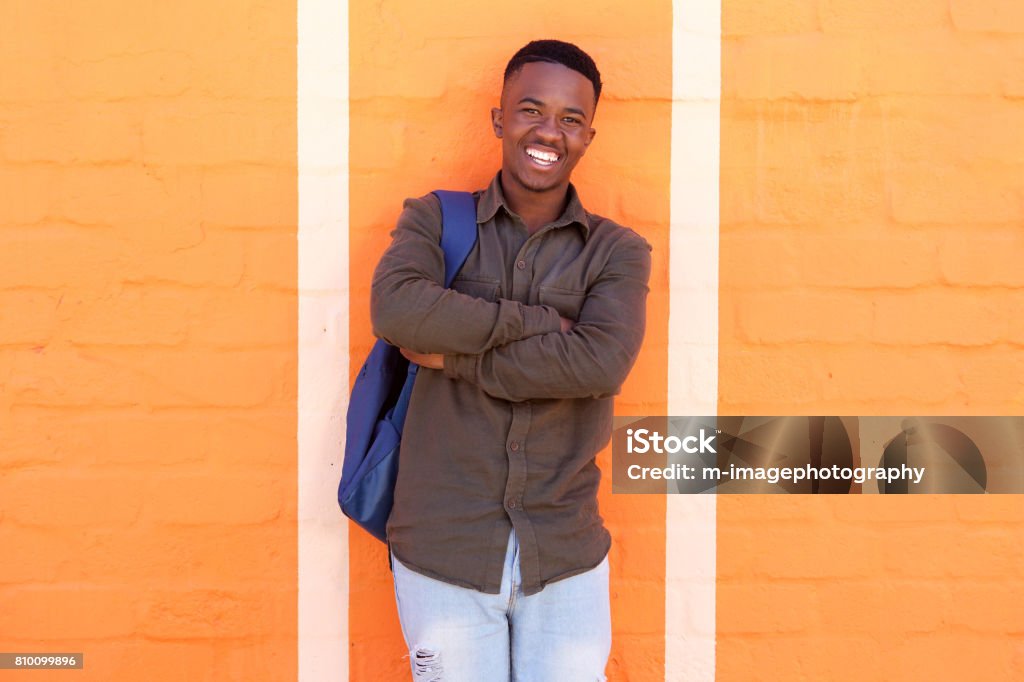 happy african male student standing against orange wall with bag Portrait of happy young african male student standing against orange wall with bag High School Student Stock Photo