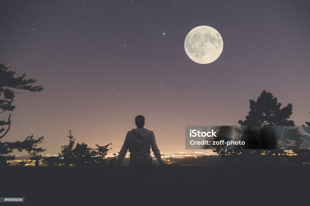 Man enjoying the view from hill above city. Full moon and stars on the sky. Young man sitting on the wall at night. City lights, moon and stars in the background. Moon Stock Photo