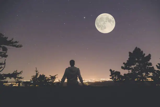 Photo of Man enjoying the view from hill above city. Full moon and stars on the sky.