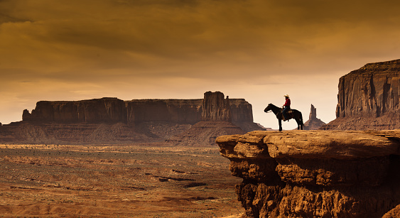 An Native American cowboy on horseback at the edge of a butte cliff at the Monument Valley Tribal Park in Arizona, USA. A famous tourist destination in the southwest USA. The iconic western landscape is a backdrop for many western movies. The native American is a Navajo tribe native. Photographed on location in desaturated sepia tone.