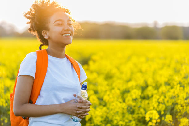 portrait en plein air de belle heureux laughing mixte jeune africaine américaine course féminine jeune femme adolescente avec bouteille d’eau potable dans un champ de fleurs jaunes au coucher du soleil sous le soleil de soirée doré - yellow flowers flash photos et images de collection