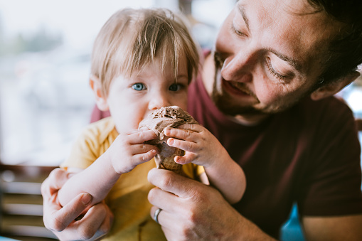 A dad feeds his one year old son a chocolate ice cream cone, the child enjoying sharing the sweet treat.  A depiction of a supportive and available father figure.