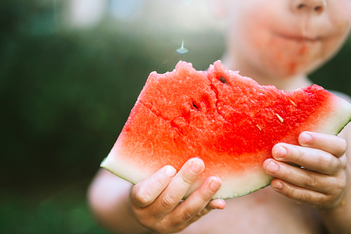 A child beats the summer heat with a juicy slice of watermelon, enjoying the sun while eating the sweet fruit.  Horizontal image with copy space.