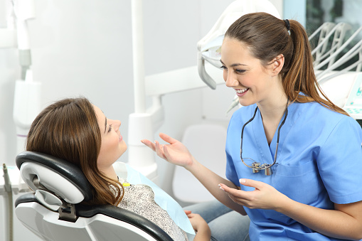 Dentist wearing blue coat explaining procedure to a patient who is sitting on a chair in a clinic box before treatment