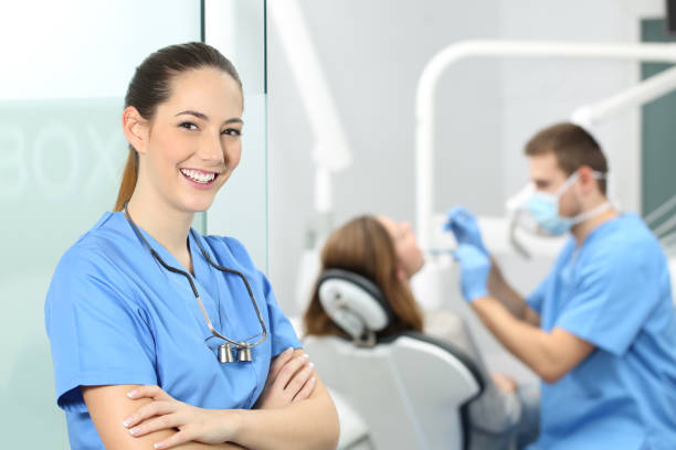 Dentist female posing at consultation Dentist female with crossed arms wearing blue coat posing and looking at you at consultation with a doctor working with a patient in the background dental hygienist stock pictures, royalty-free photos & images