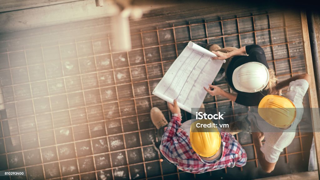 Female architect and two consruction workers on a construction site High angle view of three people with helmets, female architect, foreman and engineer on a construction site, looking down on a blueprint, copy space. Construction Site Stock Photo