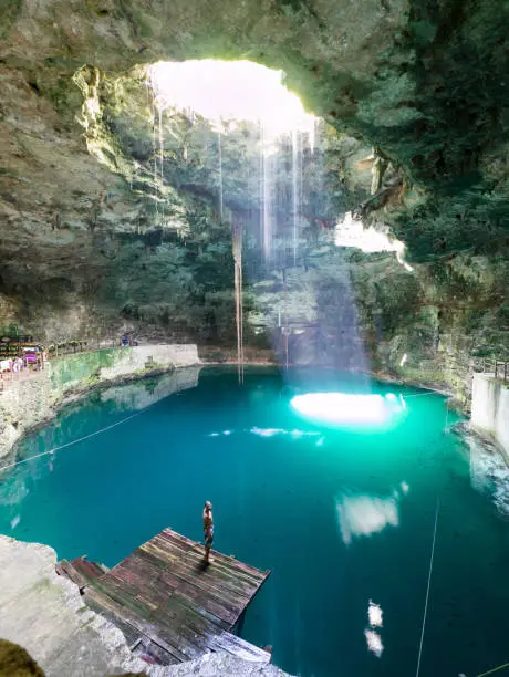 Young, white, caucasian, male man standing on wooden pier in natural pool, cave, cenote Hubiku, illuminated by light, sunbeam from above, close to Valladolid, Mexico