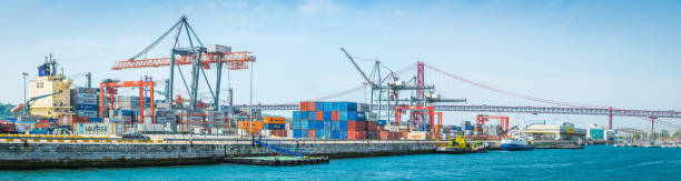 Container ship loaded with shipping containers docked in the busy cargo port at Lisbon, beneath the span of the 25 de Abril Bridge, Portugal.