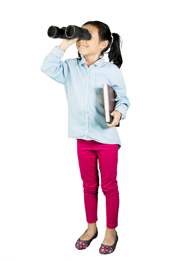 Schoolgirl looking something with binoculars while holding a textbook, isolated on white background