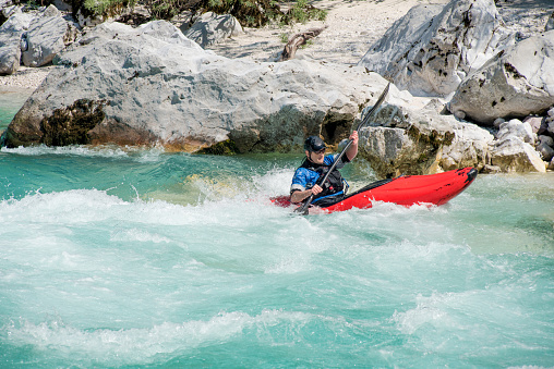 A person wearing a protective helmet and life jacket expertly navigates through turbulent whitewater rapids, with large rocks and a dense forest in the background.