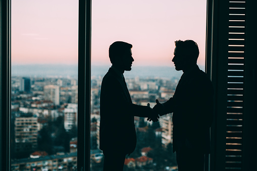 The sillouettes of two businessmen shaking hands at the window of an office.