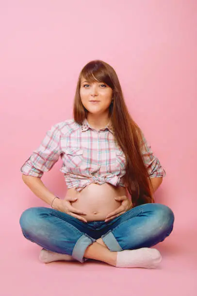 Pregnant woman in jeans and checkered shirt sitting cross legged on pink background. Happy mother-to-be. Space for text.