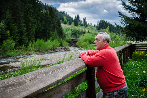 Portrait of a handsome senior man in bright sweater relying on the wooden fence near the river in the mountines and thinking . Serious senior man with gray hair and beard. Horizontal image.