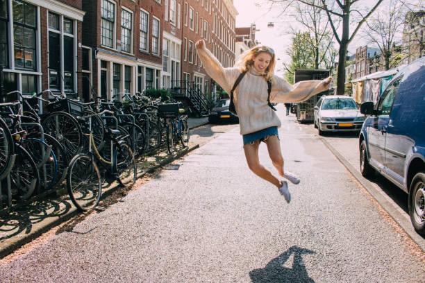 Leaping for Joy Young woman leaping into the air wearing a mini skirt and an oversized jumper. She is smiling and wearing a backpack with sunglasses on her head 21 24 months stock pictures, royalty-free photos & images