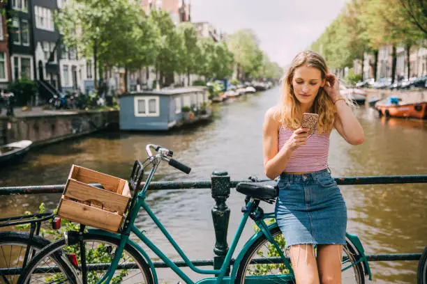 Young woman resting against a bicycle and railing at the canal while she uses her mobile telephone